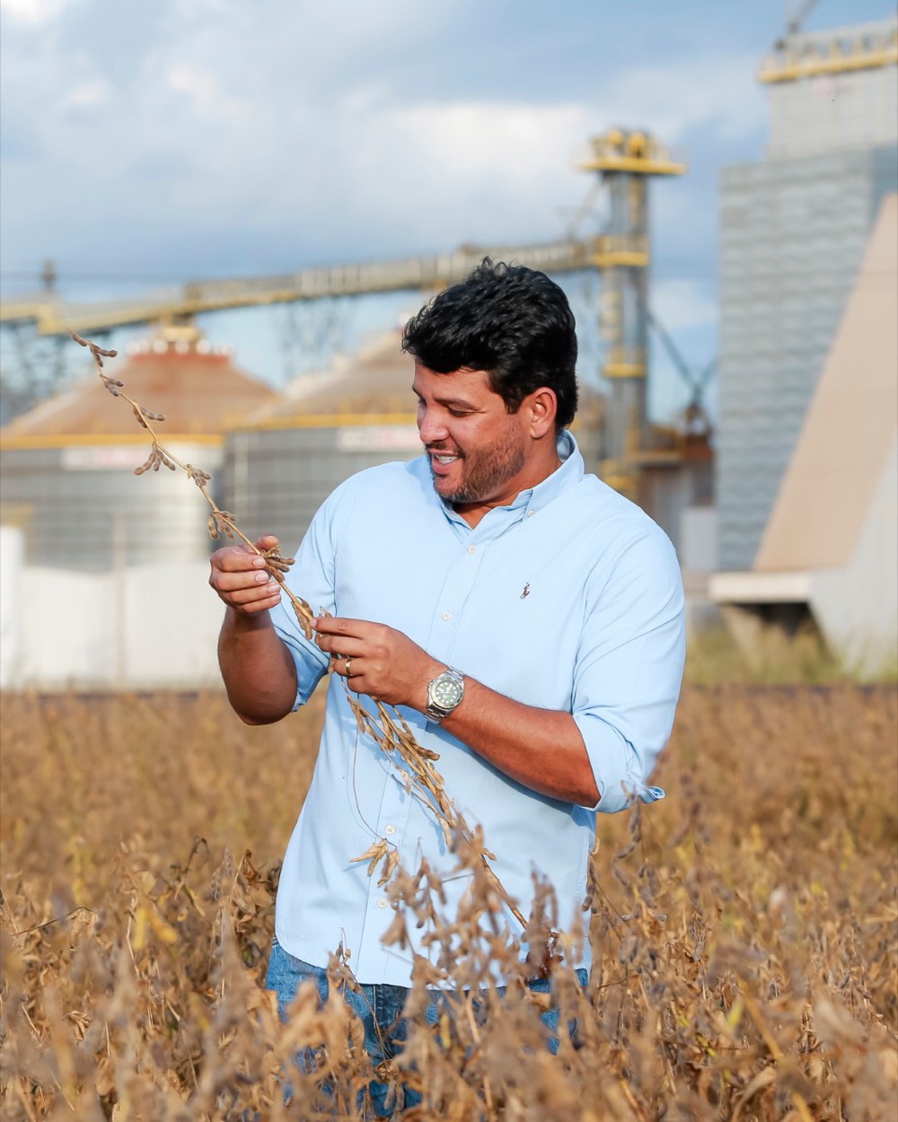 Benhur Willian segurando uma planta de soja em uma lavoura, com silos ao fundo